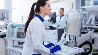 Person mit dunklen langen Haaren und weiß-blauer Schutzbekleidung sowie blauen Schutzhandschuhen bedient einen Computer in einem Labor