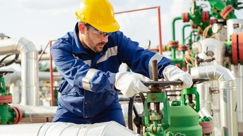 Eine Person mit Helm, Schutzbrille, Handschuhen und blauer Arbeitskleidung dreht an einem Ventil. Links und rechts davon sind verschiedene Pipelines