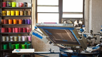 Siebdruckmaschine in Werkstatt, im Hintergrund Fenster und Regal mit Farben