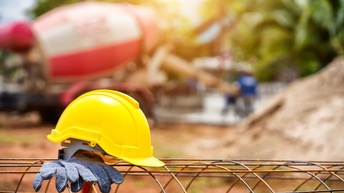 Auf einem abgerundeten Zaun aus Draht liegen ein Bauarbeiterhelm und Handschuhe. Im Hintergrund ist verschwommen eine Betonmischmaschine zu sehen