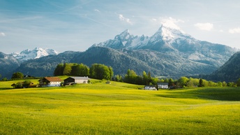 Im Vordergrund ist eine grüne Wiese mit ein paar Gebäuden sowie grünen Bäumen. Dahinter ist ein Bergmassiv mit teilweise schneebedeckten Gipfeln
