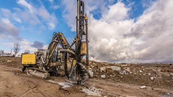 Ein Baufahrzeug mit einem massiven Bohrer bohrt eine Grube in schlammige Erde. Der Boden ist abfallend. Auf dem Boden liegen teilweise größere Steine. Am Himmel sind Wolken