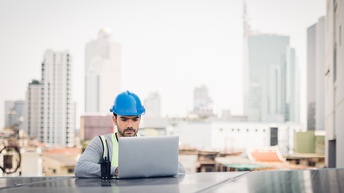 Frontale Aufnahme einer Person mit Bauhelm, die an einem Solarpanel steht und auf diesem einen aufgeklappten Laptop hat, auf dessen Bildschirm sie blickt. Im Hintergrund sind Hochhäuser