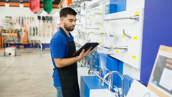 Seitliche Aufnahme einer Person mit kurzen, dunklen Haaren und Bart sowie mit Schürze, die in einem Geschäft vor einer Wand mit verschiedenen Badezimmerarmaturen steht. Die Person hält in der rechten Hand ein Tablet