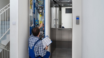 Eine Person in Arbeitskleidung hält ein Clipboard in der Hand und repariert einen Aufzug. Neben der geöffneten Lifttüre befindet sich ein elektronisches Panel, an dem die Person einen Schalter betätigt.