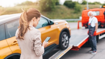Person mit geschlossenen langen Haaren blickt auf ein Smartphone, während im Hintergrund eine Person in Arbeitskleidung steht und ein Auto abschleppt