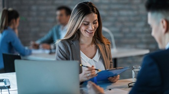 Lächelnde Person blickt auf Clipboard mit Stift in der Hand, ringsum weitere Personen und aufgeklapptes Notebook