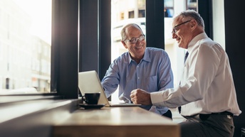 Zwei lächelnde Personen sitzen an Hochtisch an Fenstern, am Tisch aufgeklapptes Notebook und Kaffeetasse
