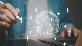 Person hält einen weißen Stift in der Hand auf dem sich eine Weltkugel mit Personen-Icons, die vernetzt sind in der Farbe weiß visualisieren, daneben steht ein Laptop auf einem Tisch