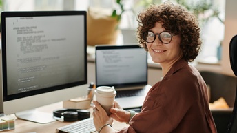 Eine zur Kamera gewandte Person mit Brille und kurzen, lockigen Haaren sitzt an einem Schreibtisch. Vor ihr steht ein Bildschirm und eine Tastatur. Rechts daneben steht ein aufgeklappter Laptop. In den Händen hält sie einen Papierbecher