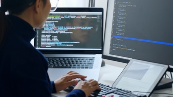 Person sitzt vor mehreren Bildschirmen und tippt auf Tastatur