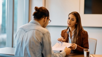Zwei Personen sitzen sich an einem Tisch gegenüber. Eine der Personen hat den Rücken zur Kamera gewandt. Die andere Person hält ihr einen Zettel hin und deutet mit einem Stift auf eine Stelle. Die andere Person blickt darauf