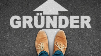 Top-Shot der hellbraunen Schuhe einer Person, die auf einem asphaltierten Boden stehen. Vor den Schuhen steht in weißer Schrift Gründer. Darüber ist ein Pfeil, der in den oberen Bildbereich zeigt