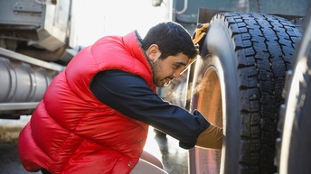 Person in roter Daunenweste hockt an großem Reifen eines Lastkraftwagens und streckt Hand in Felgenbereich, Atemluft der Person ist sichtbar