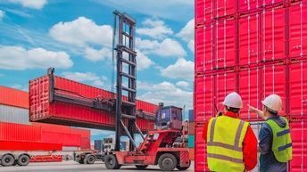 Zwei Personen mit gelben Warnwesten und weißem Schutzhelm blicken auf einen Containerstapler, der Container umsetzt, daneben stehen weitere rote Container gestapelt