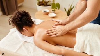 Person massiert den Rücken einer Person, die auf einer Massageliege liegt, im Hintergrund stehen Grünpflanzen