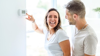Zwei Personen stehen neben einem Thermostat, das an einer Wand montiert ist. Die linke Person stellt die Temperatur am Thermostat ein. Die Personen blicken sich an