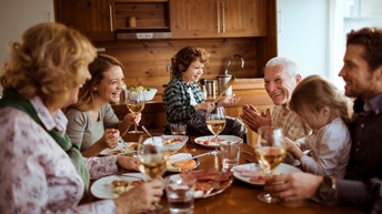 Kinder und Erwachsene sitzen um einen Tisch. Alle blicken sich gegenseitig an und interagieren miteinander. Auf dem Tisch stehen Teller mit verschiedenen Speisen. Jede Person hat einen Teller vor sich.