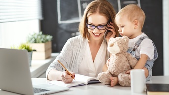 Person mit schulterlangen braunen Haaren und Brille telefoniert und macht sich daneben Notizen in ein Buch während ein Kleinkind daneben auf dem Tisch sitzt und einen Teddybär hält, am Tisch steht auch ein Laptop, eine Tasse sowie Grünpflanzen