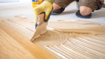 Nahaufnahme der rechten Hand einer Person, die einen Spachtel auf den Boden hält. Am Boden und am Spachtel ist eine weiße, flüssige Masse. Vor der Masse ist ein heller Holzboden. Dahinter sind die Knie einer Person auf einem hellen Boden