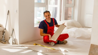 Lächelnde Person in roter Latzhose sitzt in einem Raum und verlegt einen Holzboden