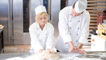 Zwei Personen in weißen Oberteilen mit Mützen stehen nebeneinander hinter einem metallenen Tisch. Vor der linken Person liegt ein großer Teig, den sie knetet sowie Mehl. Die andere Person blickt auf den Teig und ahmt mit den Händen eine Knetbewegung nach.