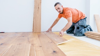 Eine Person in grauer, lockerer Hose und orangem Oberteil kniet auf dem Boden. Mit seinen Händen legt er eine Holzdiele auf den Boden, wo bereits mehrere Dielen liegen