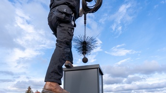 Eine Person steht auf dem Dach eines Hauses in schwarzer Kleidung. In den Händen hält er einen Kaminbesen, der gerade über der Öffnung eines Kamins schwebt