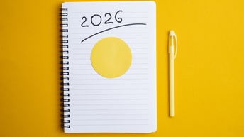 Auf gelbem Hintergrund ist ein Notizbuch, auf dem 2026 steht. Daneben liegt ein Kugelschreiber