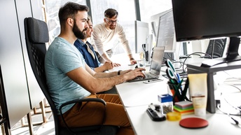 Personen sitzen bei einem Schreibtisch mit Laptops und Monitore in einem Büro.