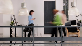 Büro mit Schreibtischen und drei Personen, die in Bewegungsunschärfe durchschreiten