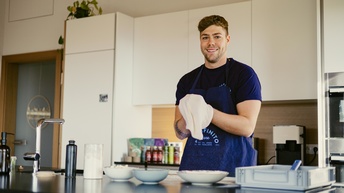 Eine Person mit Schürze steht in der Küche und trocknet mit einem Geschirrtuch ab. Sie steht hinter einer dunklen Arbeitsfläche, auf der Teller, ein Gestell mit Geschirr und ein Wasserhahn sind. Im Hintergrund sind weiße Küchenkästen und eine Tür.
