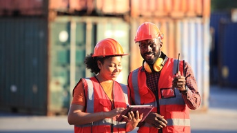 Zwei lächelnde Personen in roter Schutzkleidung mit Schutzhelm und Tablet sowie Funkgerät, im Hintergrund stehen Container in der Unschärfe