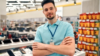 Porträt einer Person mit kurzen, dunklen Haaren, Bart, Ausweisschild um den Hals sowie verschränkten Armen, die in einem Elektrogeschäft steht