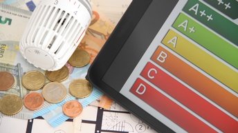 Nahaufnahme und Top-Shot eines weißen Zettels, auf dem Münzen, Geldscheine, ein Regler eines Heizkörpers und ein Tablet liegen. Auf dem Tablet sind sieben horizontale Streifen in den Farben rot bis grün. Auf dem obersten steht A+++.