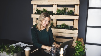 Eine Person mit hellen, offenen Haaren sitzt an einem dunklen Schreibtisch und tippt auf einer Tastatur eines Laptops. Auf dem Schreibtisch stehen kleine Topfpflanzen. An der Wand hinter der Person sind weitere Pflanzen