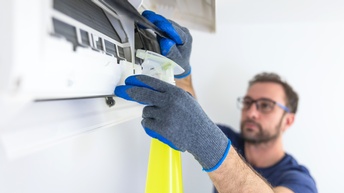 Person mit Brille und Arbeitshandschuhen hält gelbe Sprühflasche auf eine Klimaanlage, Hände und Sprühflasche sind im Bildfokus