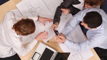 Personen sitzen rund um einen Tisch auf dem Grundrisse ausgebreitet sind und halten einen Taschenrechner, ein Laptop steht ebenso auf dem Tisch, Vogelperspektive