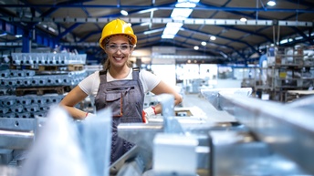 Lächelnde Person in Arbeitskleidung mit gelben Schutzhelm und Schutzbrille steht in einer industriellen Produktionsstätte