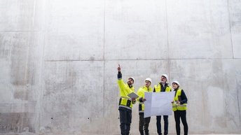 Vier Personen in gelben Warnwesten mit weißen Schutzhelmen stehen vor Betonwand und blicken nach oben in Fingerzeigrichtung einer der Personen, zwei der Personen halten großen Papierplan