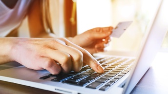 Detailaufnahme einer Person, die mit dem rechten Zeigefinger auf eine Buchstabentaste eines Laptops tippt während in der linken Hand eine Karte gehalten wird.