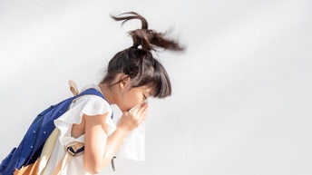 Seitliche Aufnahme eines Kindes mit Rucksack, das vor einer hellen Wand steht und sich in ein Taschentuch schnäuzt