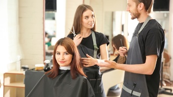 Sitzende Person mit schwarzem Haarschneideumhang um den Körper lächelt sanft, hinter ihr zwei weitere Personen in Schürzen mit Kamm und Schere in Unterhaltung