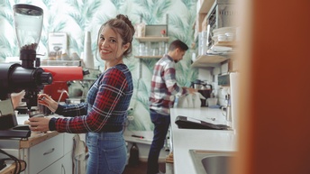 Lächelnde Person in karierter Jacke steht an Kaffeemühle im Barbereich eines Lokals, im Hintergrund weitere Person
