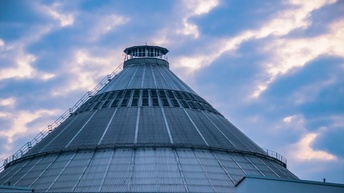Außenansicht eines großen zylindrischen, metallischen Gebäudes zur Abfallbehandlung vor einem bewölkten Himmel
