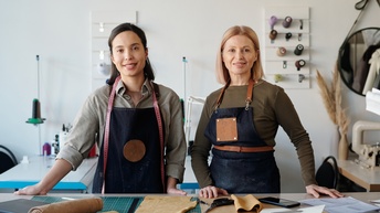 Zwei Personen in Schürzen stehen an einem Tisch. Vor ihnen liegen Scheren, Stifte, Zangen, Notizhefte, Stoffrollen aus Leder und Lederstücke. Die linke Person hat ein Maßband um den Hals