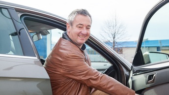 Freudige Person mit Jacke sitzt auf dem Beifahrersitz eines Personenwagens und hält die Autotür offen