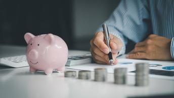 Auf einer Oberfläche sind fünf, unterschiedlich hohe Münzstapel. Links daneben steht eine kleine Figur eines Schweins. Links neben dem Schwein ist ein Taschenrechner. Dahinter sind die Arme einer Person. Unter der rechten Hand sind bedruckte Zettel