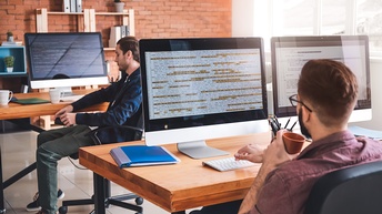 In einem Büro sitzen zwei Personen an zwei Schreibtischen. Vor der einen Person sind zwei Bildschirme, vor der anderen Person ist ein Bildschirm. Auf allen Bildschirme sind verschiedene Quellcodes