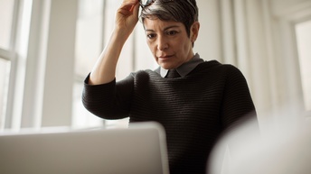 Person mit kurzen Haaren mittleren Alters steht in einem hellen Raum und hält eine Brille am Kopf während auf einen Bildschirm geblickt wird